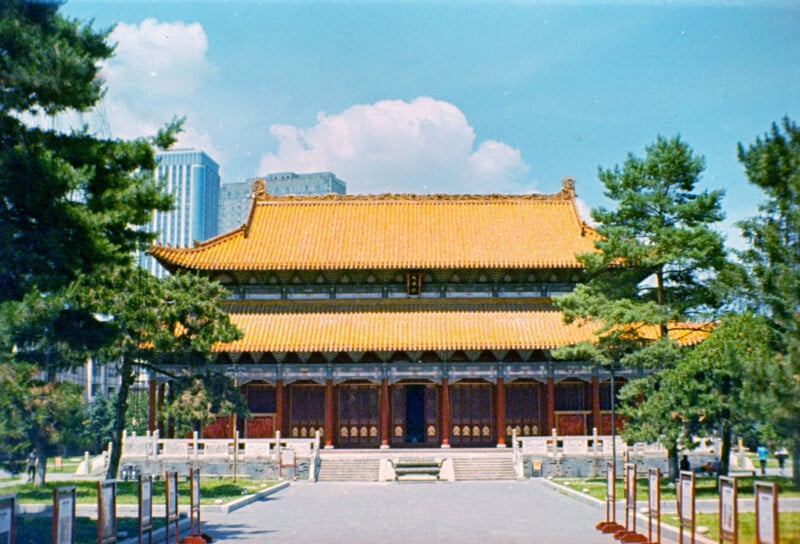 A traditional Chinese building with an ornate golden roof stands among green trees, with modern skyscrapers visible in the background under a blue sky with clouds.