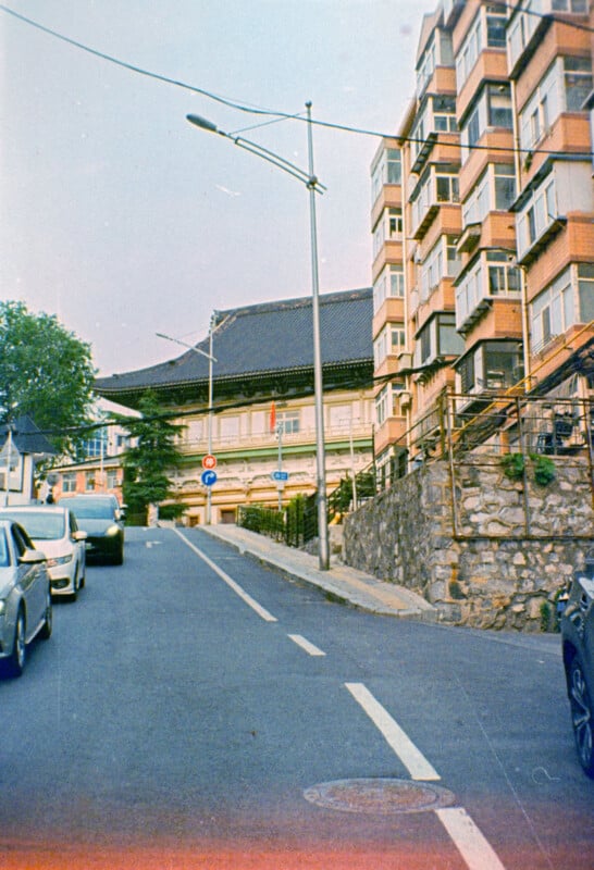 A city street slopes uphill with parked cars on the left, a modern apartment building on the right, and a traditional building with a tiled roof in the background under a clear sky.