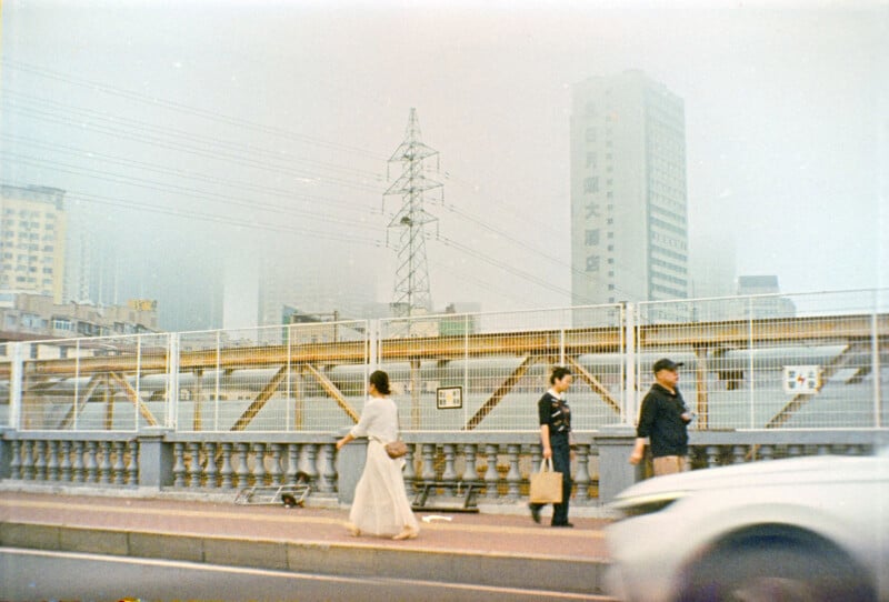 Three people walk along a city sidewalk beside a road, with a white car passing by. In the background are tall buildings, power lines, and a metal fence, all seen through a hazy sky.