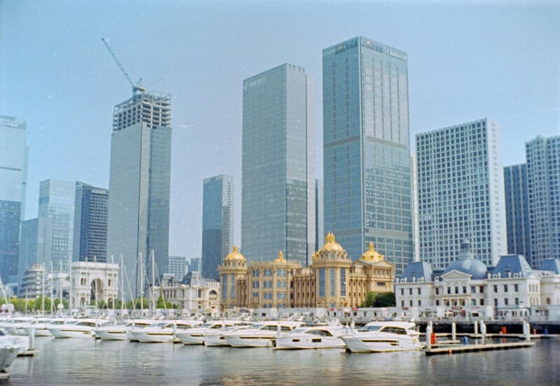 A marina with rows of white yachts sits in front of tall modern skyscrapers and an ornate golden building under a clear blue sky. A crane is visible atop one of the high-rises.