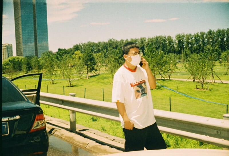 A man wearing a mask and glasses stands by a guardrail near a parked black car, holding a phone to his ear. A tall building and green park with trees are visible in the background under a partly cloudy sky.