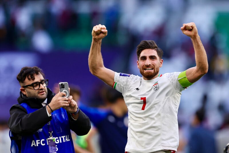 A soccer player in a white jersey with number 7 raises both fists in celebration while a media person in a blue vest takes his photo with a phone. They are on a soccer field with blurred fans in the background.