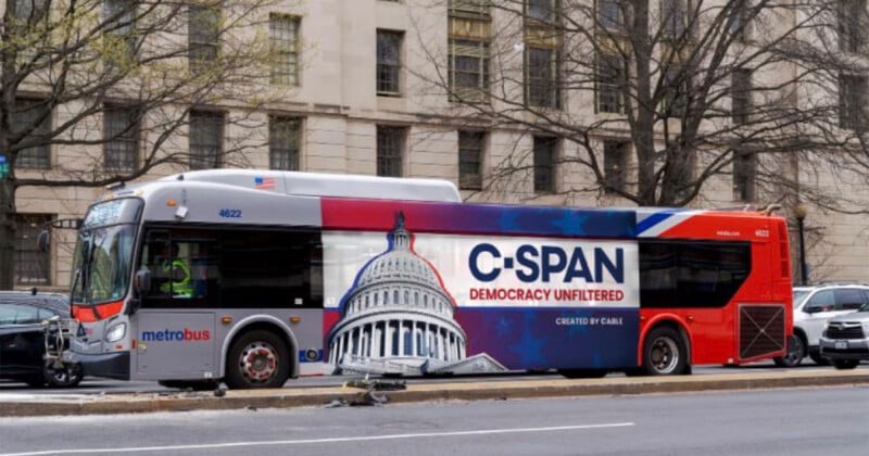 A Metrobus wrapped in a C-SPAN advertisement, featuring an image of the U.S. Capitol dome and the slogan "Democracy Unfiltered," is parked on a city street with trees and a large building in the background.