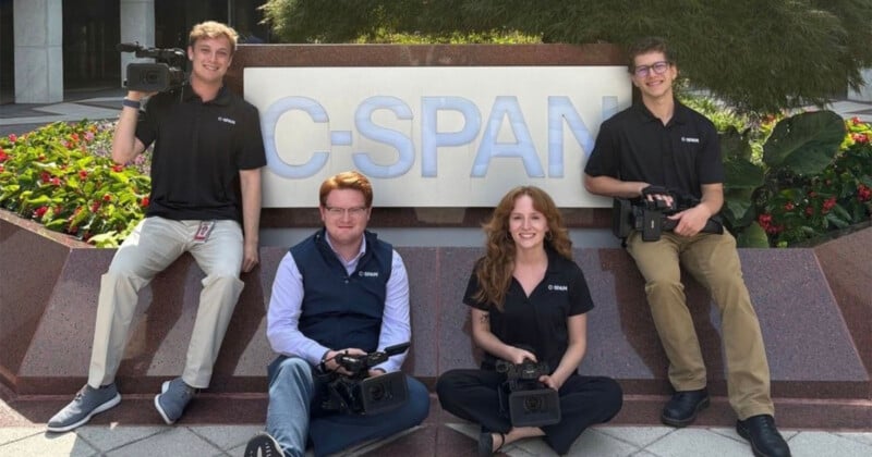 Four people wearing black C-SPAN shirts sit and kneel in front of a large C-SPAN sign outdoors, each holding a video camera, with greenery and flowers in the background.