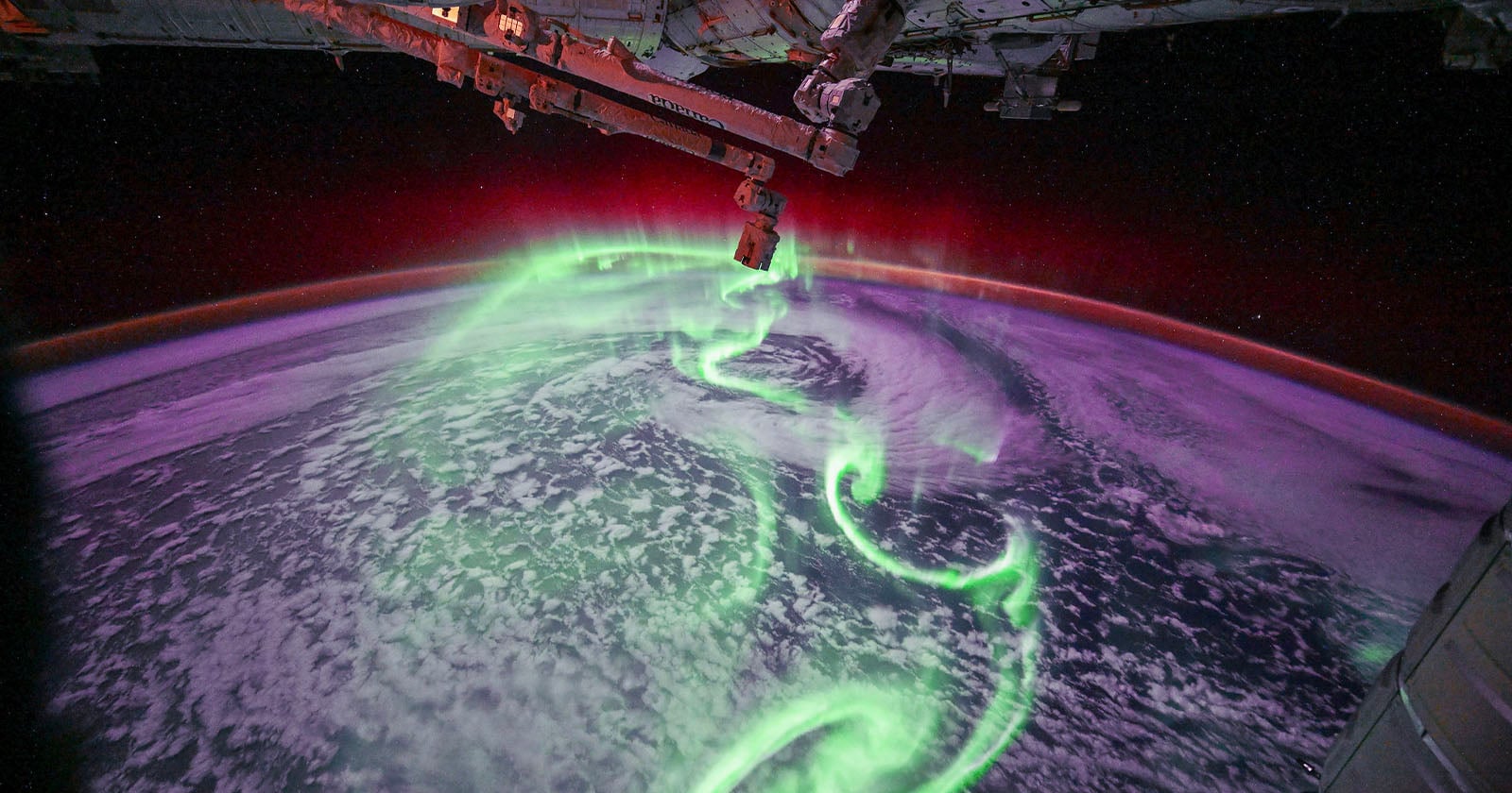 View from space showing swirling green and purple aurora lights over Earth's surface, with part of a spacecraft structure visible at the top and right edges of the image.
