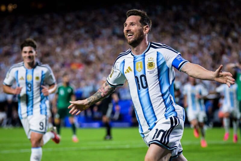 A soccer player in an Argentina jersey celebrates with arms outstretched on the field, smiling, as a teammate runs behind him. The stadium is packed with cheering fans in the background.
