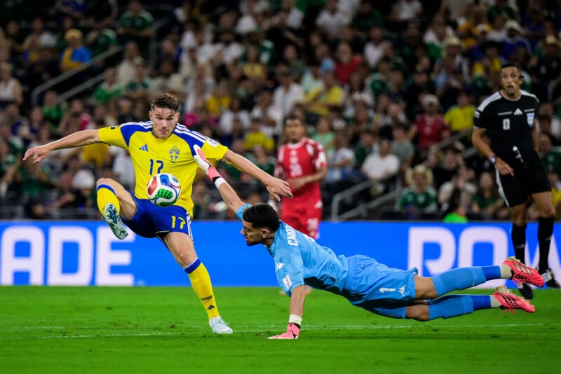 A soccer player in a yellow and blue uniform tries to kick the ball as a goalkeeper in blue dives to stop him during a match, with spectators in the background.
