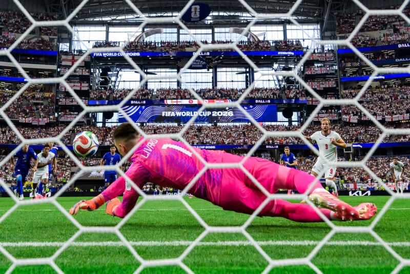A soccer goalkeeper in pink dives to stop a penalty kick as the ball heads for the goal. The crowded stadium and the net are visible, with players and fans intently watching the action at the FIFA World Cup 2026.