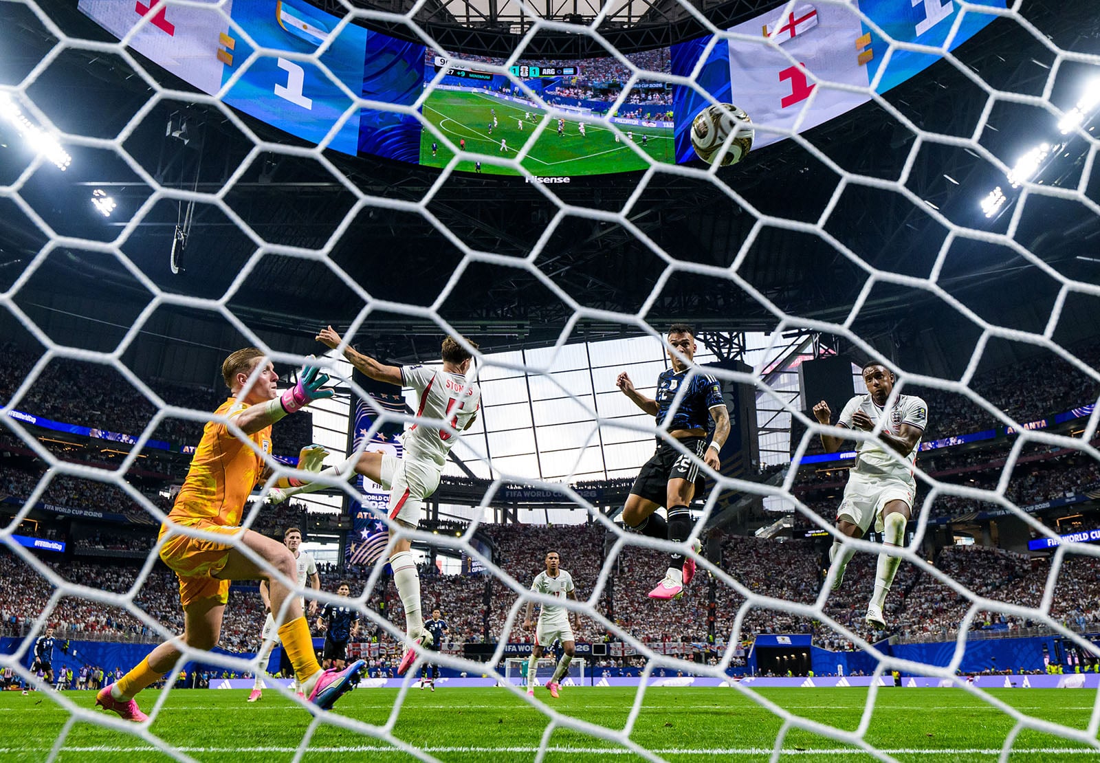 A soccer match seen from behind the goal, with players mid-air attempting to score, a goalkeeper in orange reaching for the ball, and a stadium full of spectators in the background.