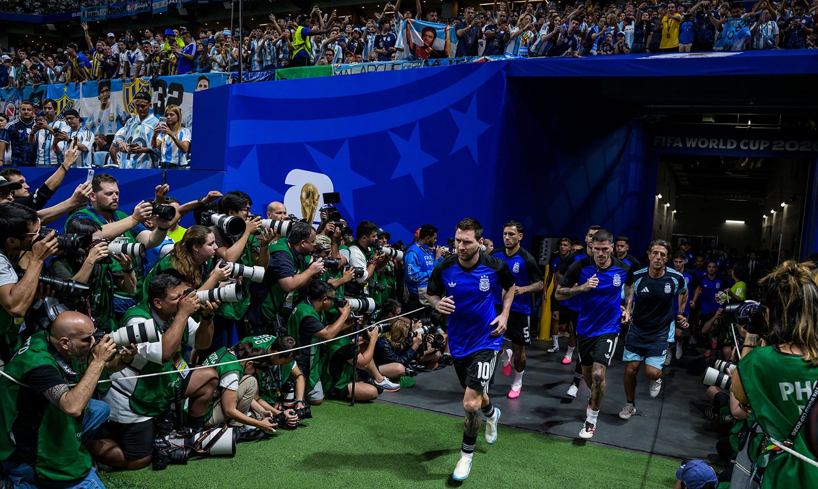 Soccer players run onto the field surrounded by photographers and fans, with a stadium crowd and FIFA World Cup signage visible in the background.