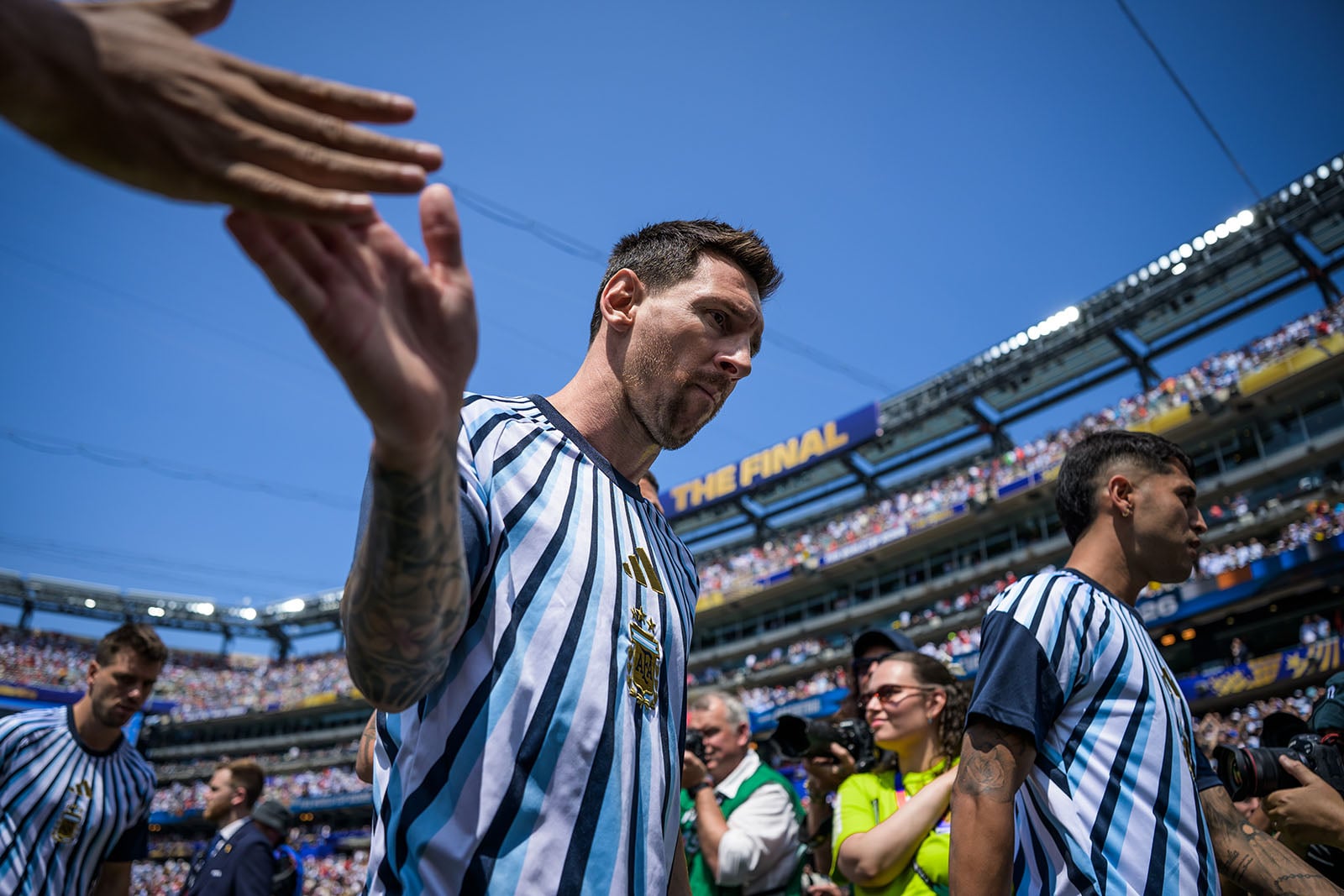 A soccer player in a striped jersey high-fives fans as he walks onto a sunny stadium field, with photographers, teammates, and a crowd in the background.