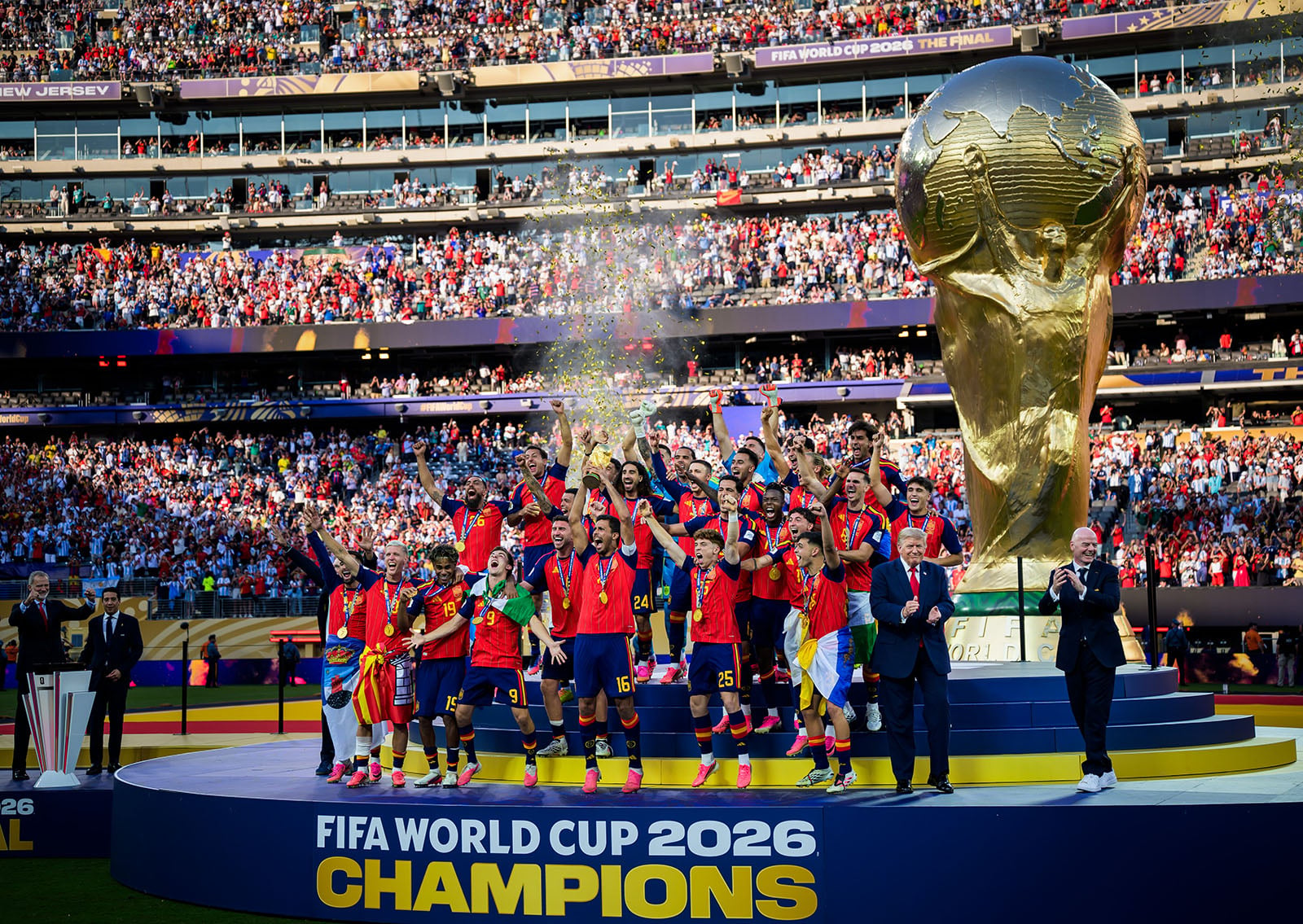 A soccer team celebrates on a podium with confetti, holding a trophy by a giant FIFA World Cup replica. The banner reads "FIFA World Cup 2026 Champions." A packed stadium is visible in the background.