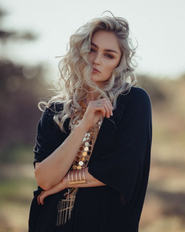 A woman with long, wavy blonde hair wearing a dark outfit and gold jewelry stands outdoors, looking into the camera with a serious expression. The background is softly blurred.