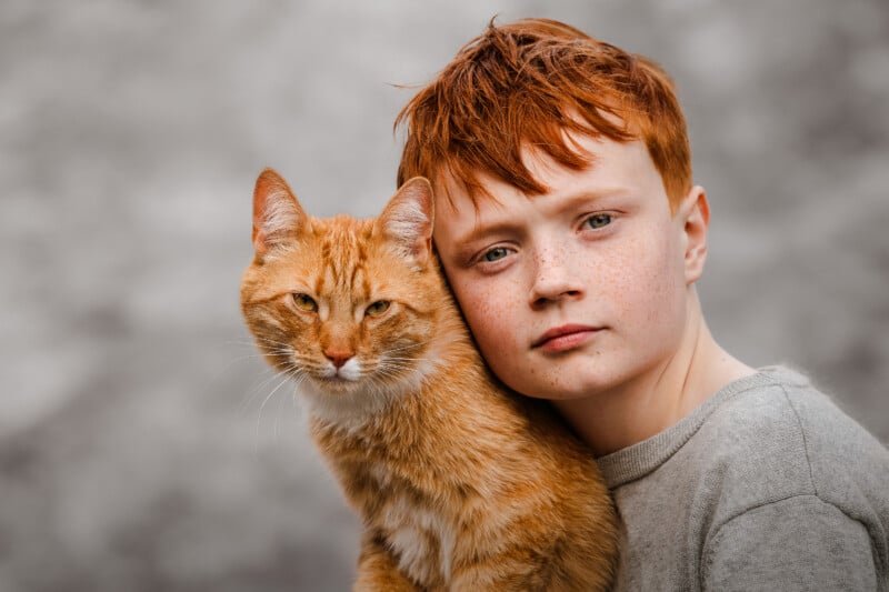 A red-haired boy with freckles wearing a gray shirt poses closely with an orange tabby cat, both looking directly at the camera against a neutral, blurred background.