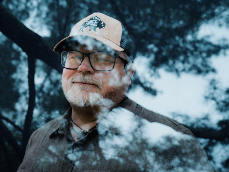 A middle-aged man with glasses, a gray beard, and a cap stands outdoors, partially reflected in glass. Trees and sky are visible in the background and foreground, creating a layered, dreamy effect.