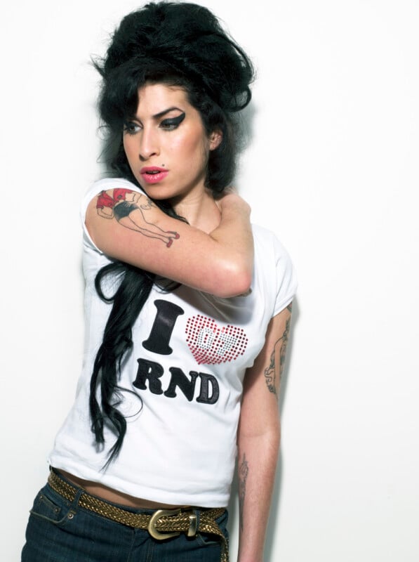 Amy Winehouse with her signature beehive hairstyle and winged eyeliner wears a white t-shirt with an "I heart RND" graphic and a gold belt.