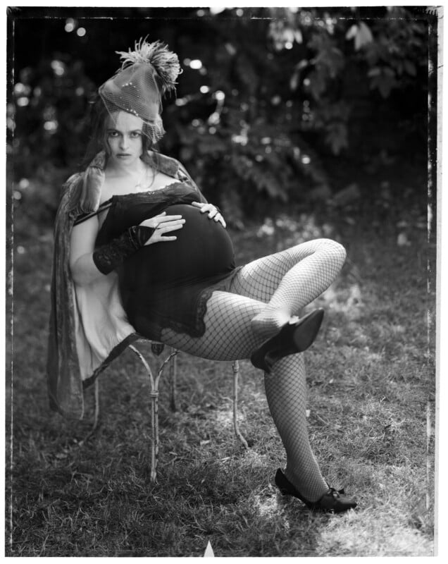 A pregnant woman with dark hair, wearing a lace-trimmed slip, fishnet stockings, and a feathered fascinator, sits on a chair outdoors.