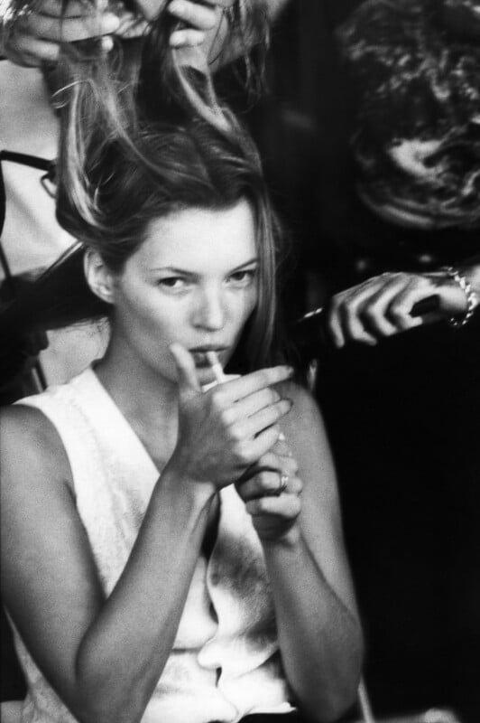 A black-and-white close-up of Kate Moss lighting a cigarette while a stylist works on her hair.