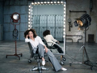 Sandra Oh sits in a salon chair in front of a large, illuminated vanity mirror, flanked by two large studio lights in an industrial space.