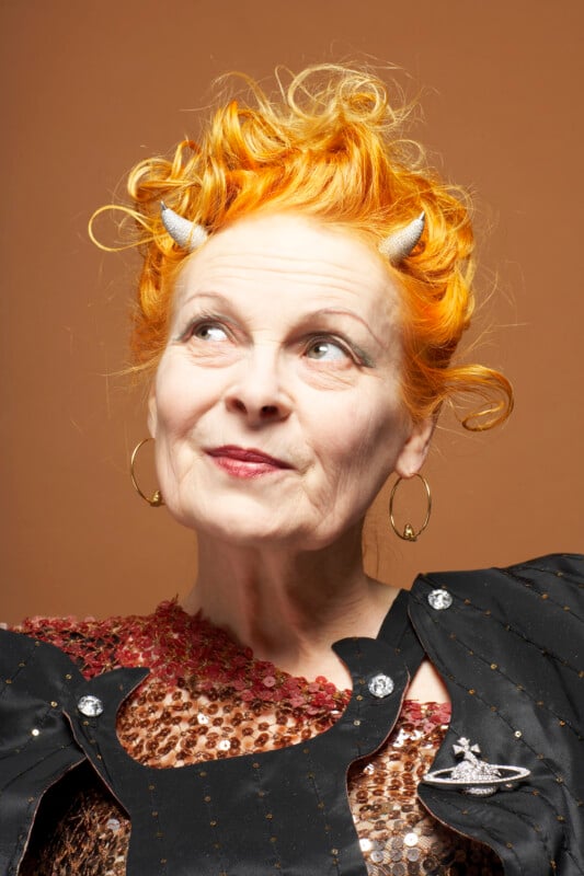 A portrait of Vivienne Westwood with bright orange hair styled with two small silver horns, wearing gold hoop earrings and a black jacket over a sequined top.
