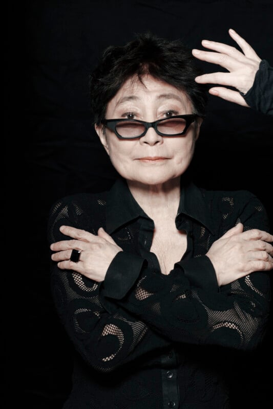 Yoko Ono poses against a black background, wearing black glasses and a black patterned shirt with her arms crossed.