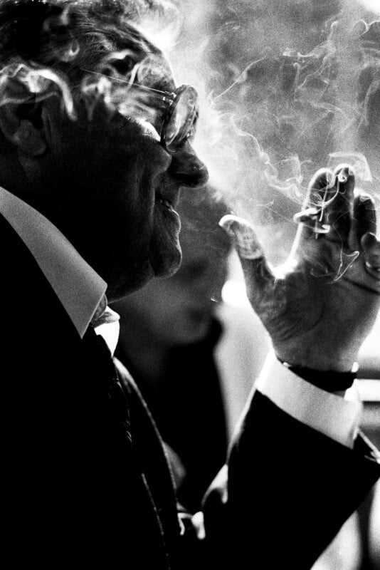 A man in a suit and glasses smokes a cigarette, with swirling smoke obscuring part of his face. The black and white image captures him in profile, smiling slightly.