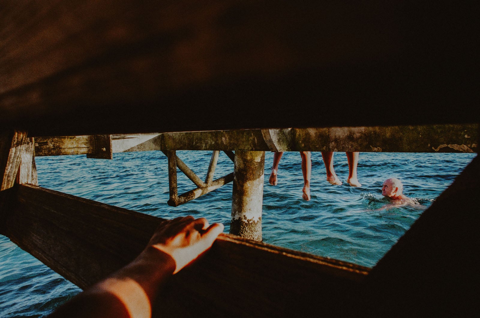 A person’s hand grips a wooden railing, looking through a gap at people sitting on a pier with their legs dangling over blue water, while someone swims below.