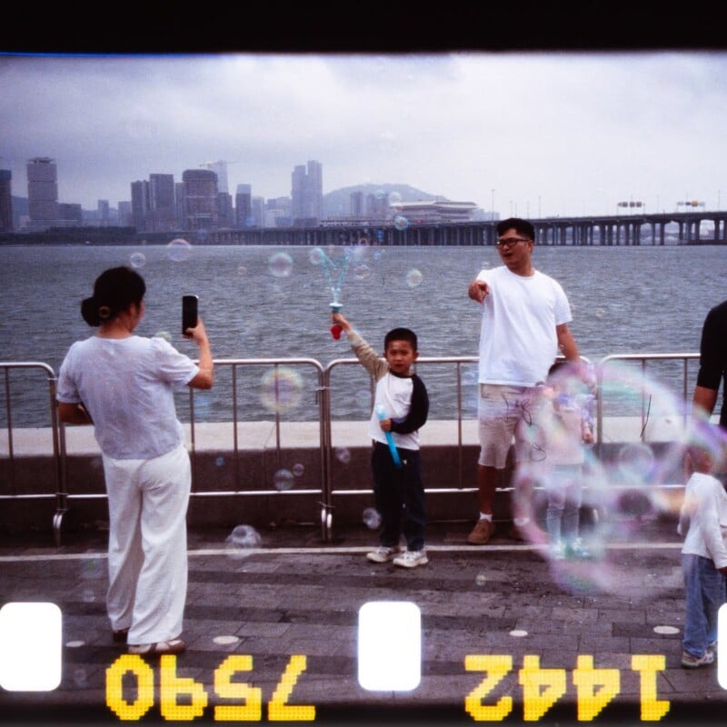 A child holding a bubble wand poses while a woman takes a photo on a waterfront promenade. Two men stand nearby. City buildings and a bridge are visible across the water under a cloudy sky. Film strip edges frame the image.