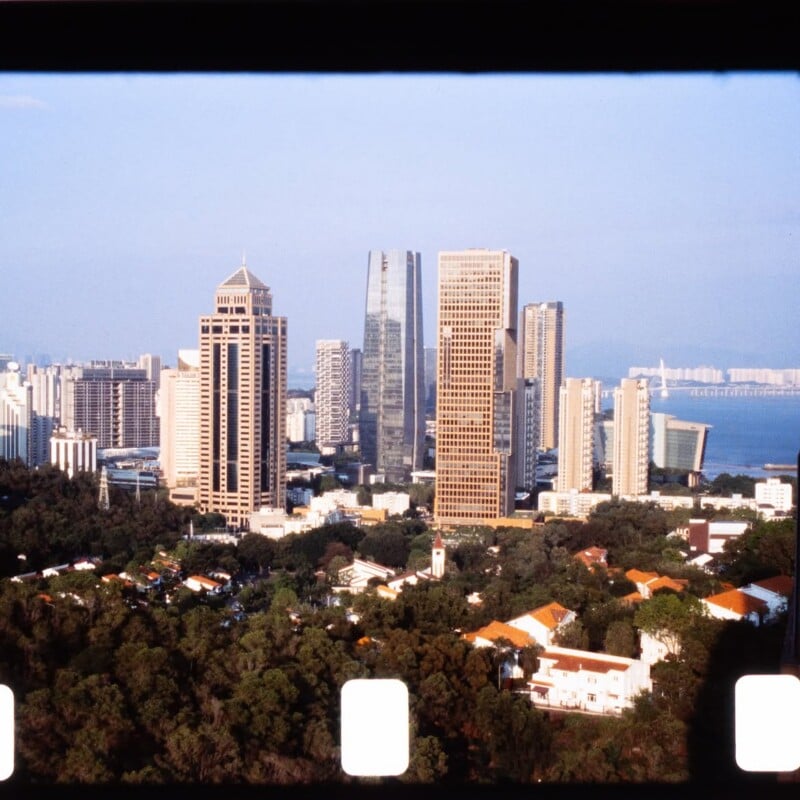 A cityscape featuring tall modern skyscrapers, a mix of older buildings with orange rooftops, and lush green trees in the foreground, viewed through a film frame with visible sprocket holes.