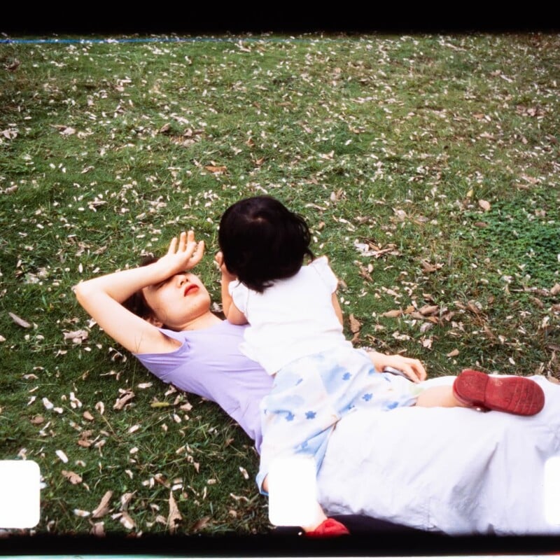 A woman lies on the grass with her arm over her forehead, while a young child sits on her, facing her. The scene is outdoors, with scattered leaves on the ground.