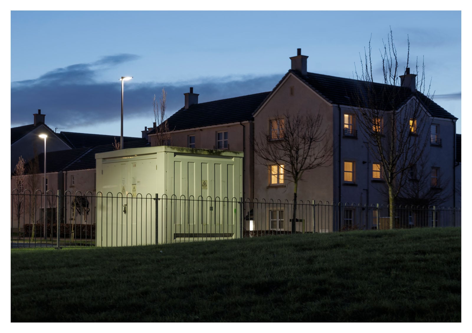 A small utility building surrounded by a metal fence is lit by streetlights at dusk, with two residential buildings and leafless trees in the background. Some apartment windows are warmly illuminated.