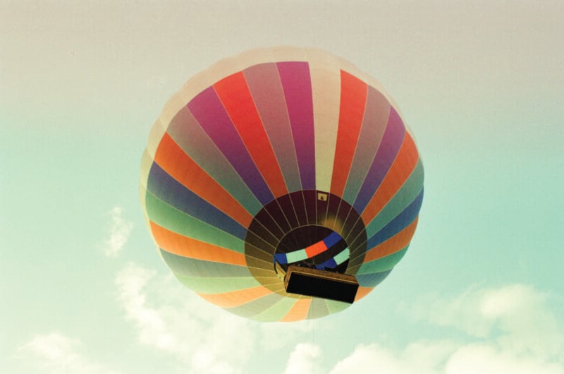 A colorful hot air balloon with red, blue, green, and orange panels floats in the sky, viewed from below against a light, slightly cloudy background.