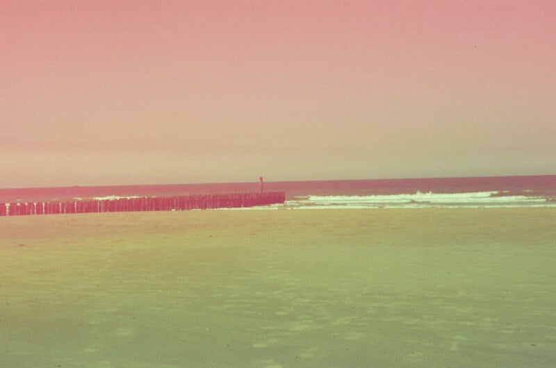 A sandy beach under a pink-tinted sky, with gentle waves and a wooden breakwater extending into the water.