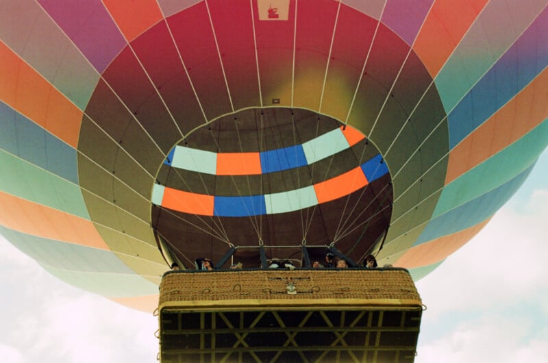 A view from below of a colorful hot air balloon with people looking out from the basket as it floats in the sky.