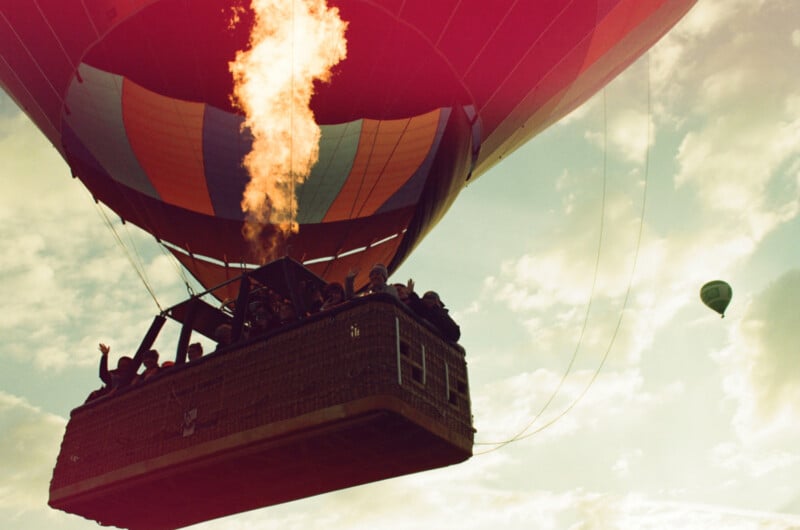 A large hot air balloon rises, its flame burning brightly, with people in the basket looking out. Another small balloon is visible in the sky against a backdrop of clouds.