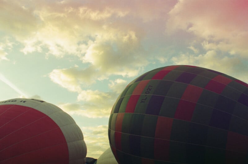 Two partially inflated hot air balloons, one red-and-white and one with dark green and red stripes, set against a cloudy sky during what appears to be early morning or late afternoon.
