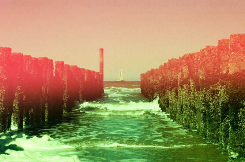 A sailboat on the horizon is seen between two mossy sea walls with waves crashing. The image has a reddish-pink haze or filter, giving it a vintage feel.