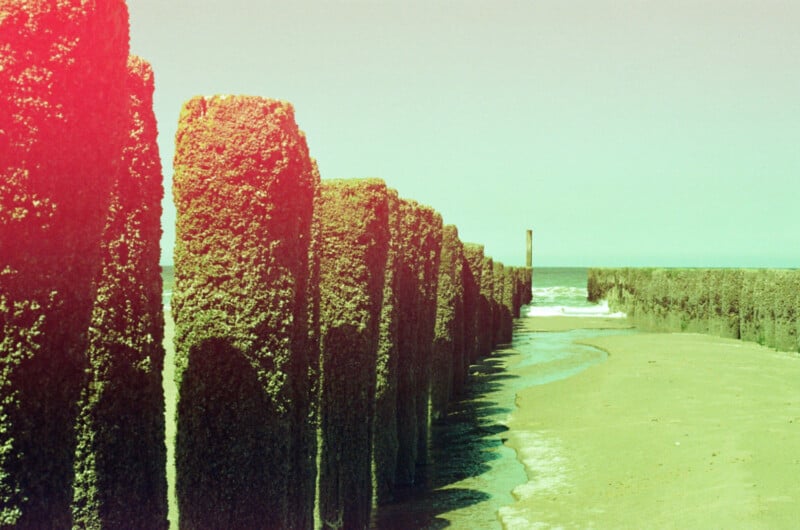 Rows of moss-covered wooden posts line a sandy beach, casting shadows. The sea and distant horizon are visible, and the image has a red light leak effect on the left side.