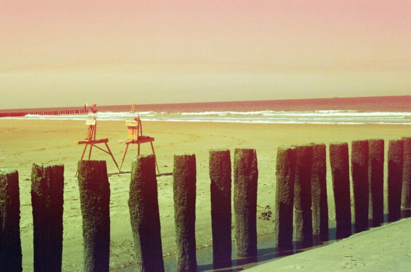 Two wooden easels stand on a sandy beach near the shoreline, behind a row of algae-covered wooden posts, with waves and a pastel sky in the background.