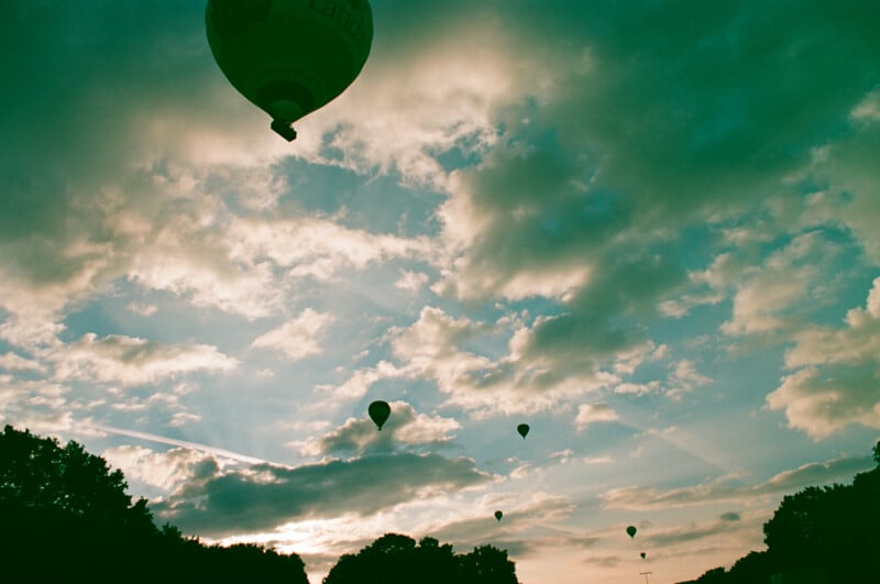 Several hot air balloons float across a cloudy sky at sunset, silhouetted against the light. Dark trees line the bottom of the image.