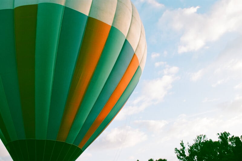 A close-up of a colorful hot air balloon with green and orange panels floats against a blue sky with scattered clouds. Tree tops are visible in the lower right corner.