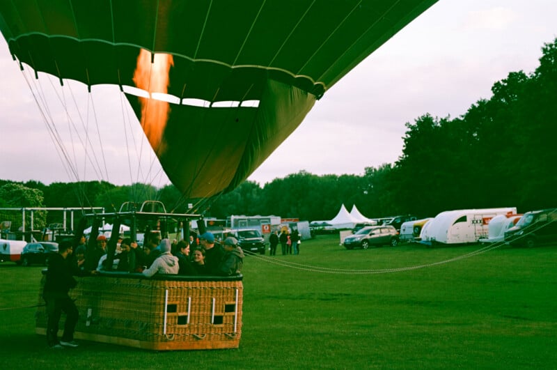 A hot air balloon is being inflated with a visible flame, while people sit in the basket. There are cars, tents, and people in the background on a grassy field.