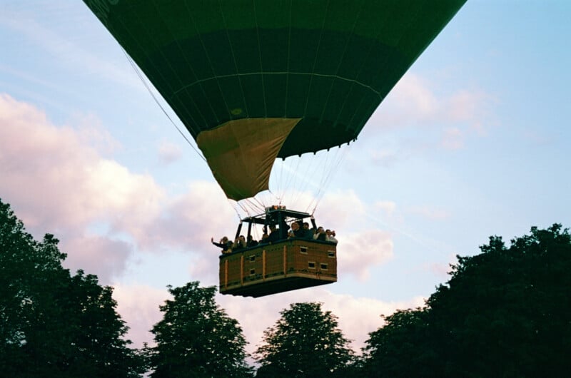 A hot air balloon carrying people in a wicker basket floats above treetops against a blue sky with clouds, viewed from below.