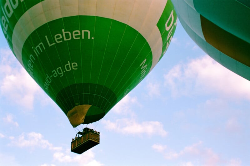 Green and white hot air balloon with people in the basket floats in the sky, with parts of other balloons visible nearby. Blue sky and scattered clouds are in the background.