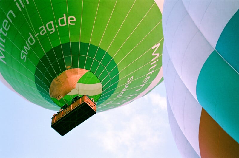 Close-up view of colorful hot air balloons ascending into the sky, with a green balloon and basket prominently visible, and portions of other balloons alongside.