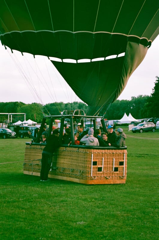 A group of people sits in a hot air balloon basket on a grassy field, preparing for takeoff. The balloon canopy is partially inflated above them. Cars and tents are visible in the background.