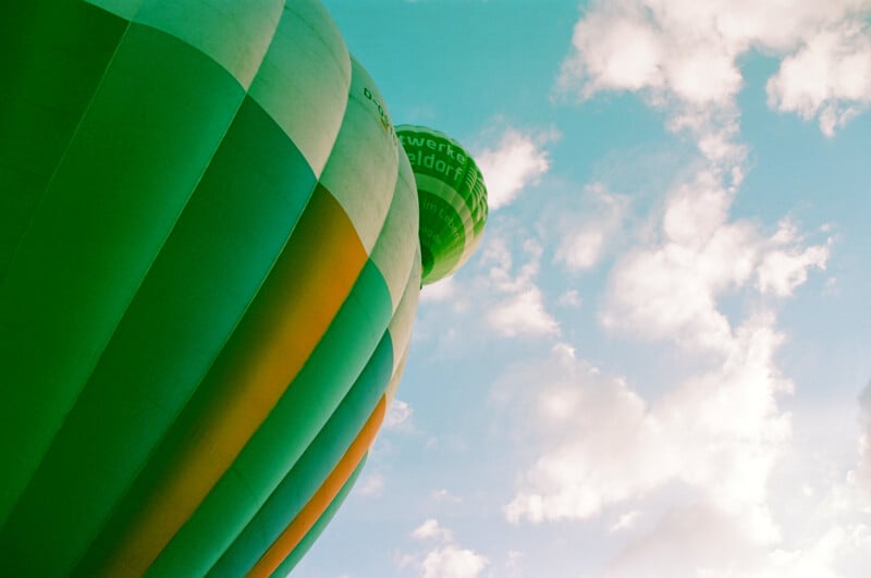 A close-up of a green and yellow hot air balloon rising against a partly cloudy blue sky, with part of another green balloon visible in the background.