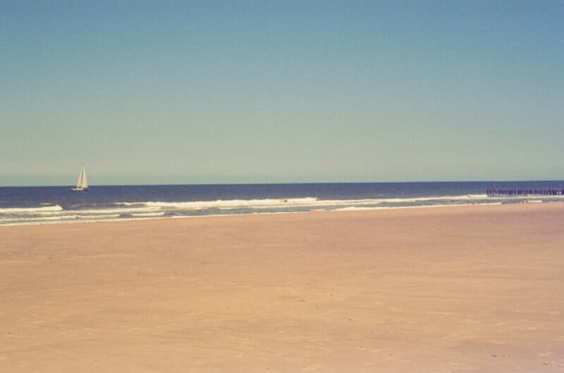 A sailboat glides on the ocean under a clear blue sky, with gentle waves meeting a wide, empty sandy beach in the foreground.