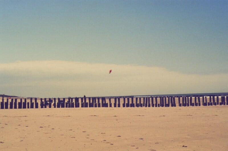 A sandy beach with wooden posts aligned in a row and a single red kite flying in the blue sky above the horizon.