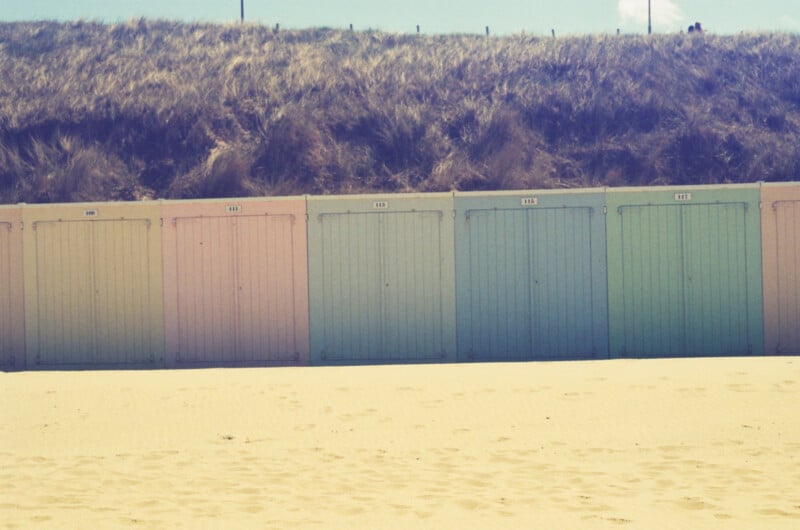 Colorful pastel beach huts with closed doors stand in a row on sand, set against a backdrop of grassy dunes under a bright, sunny sky.